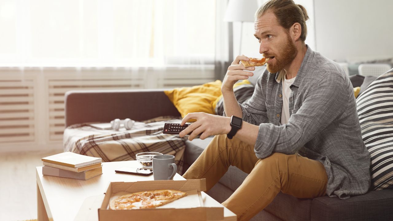 A man sitting on a couch, holding a remote, eating pizza, watches TV, pizza box, books rest, relaxation