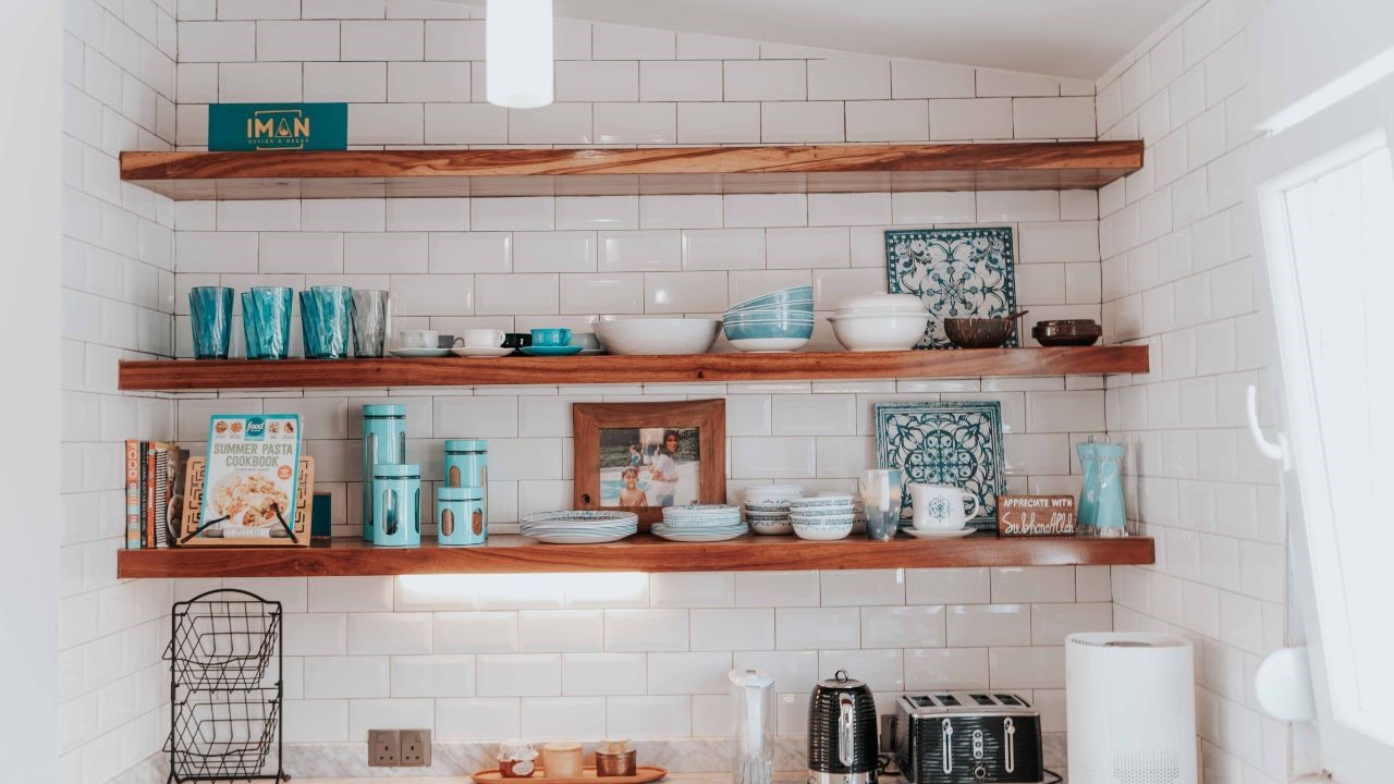 A neatly organized kitchen with wooden floating shelves holding blue and white dishes, glassware, cookbooks, and decorative items, against a backdrop of white subway tile.