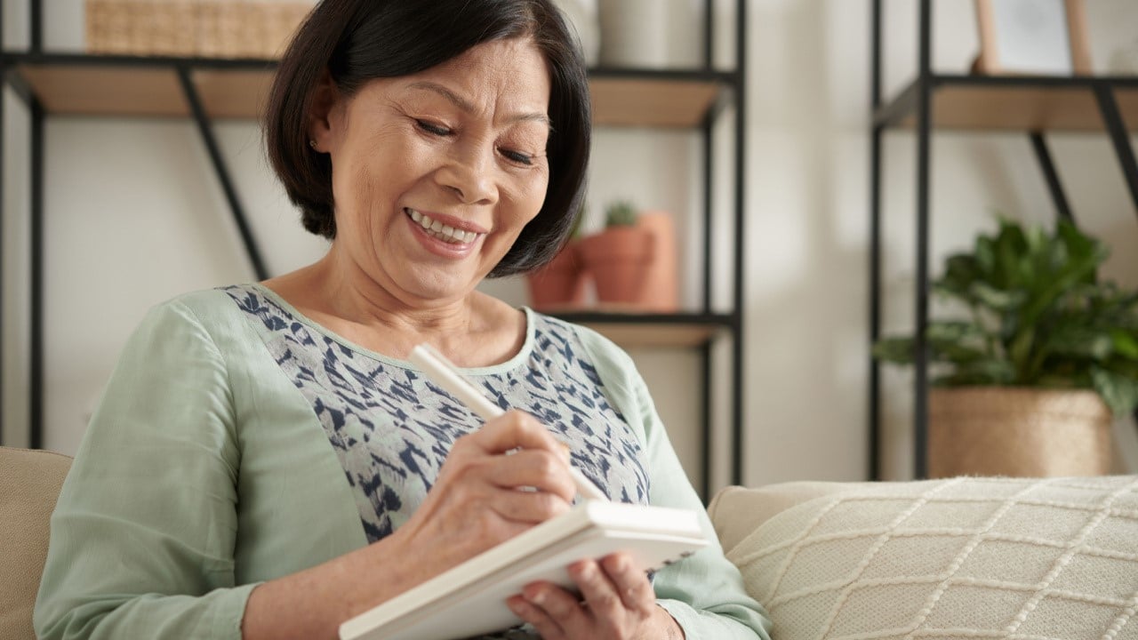 A smiling elderly woman writing thoughts in gratitude journal as a part of self care and new year resolution