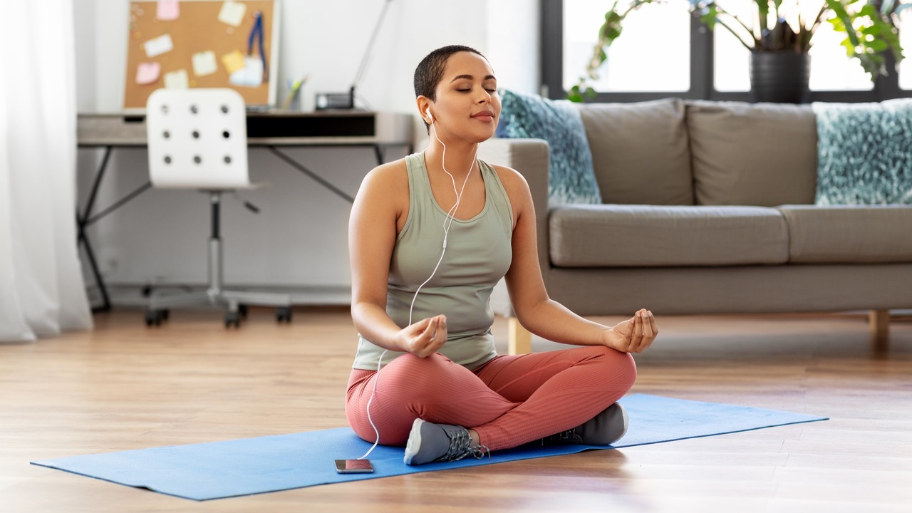 A woman sitting on a yoga mat in her living room, listening to something as she meditates.