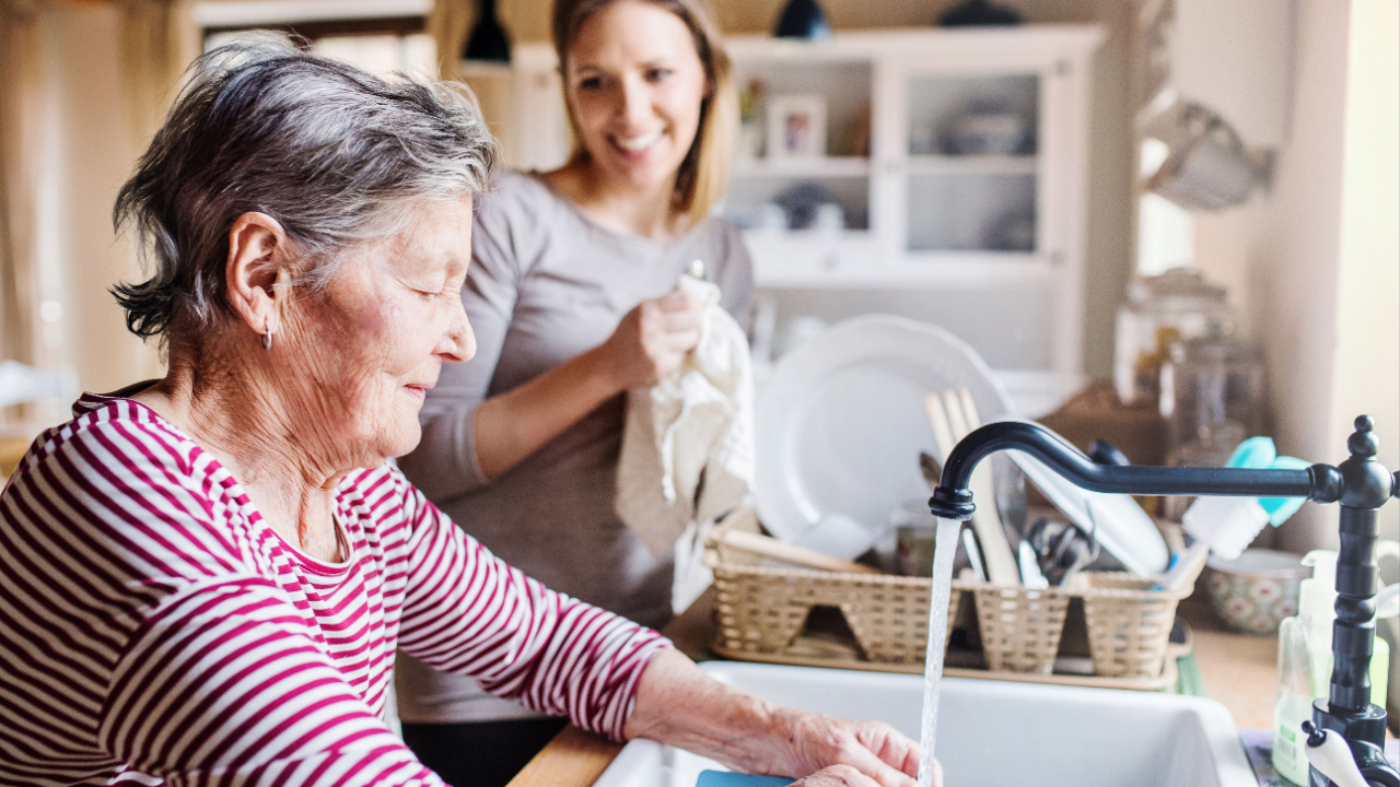 An elderly grandmother with an adult granddaughter at home, washing the dishes in running water