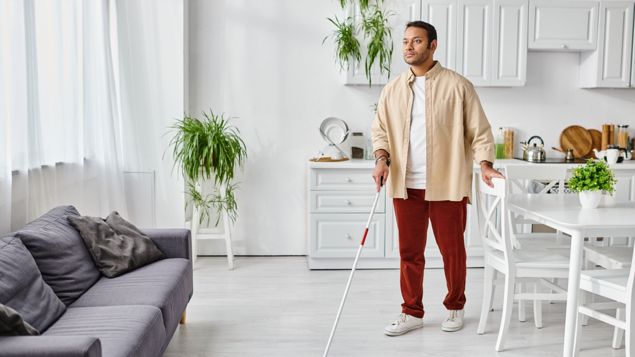 Blind man in casual cozy homewear using walking stick at home between furniture, disabled person