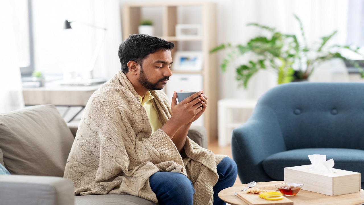 Cold, sick young man in blanket drinking hot tea with lemon, honey and ginger at home