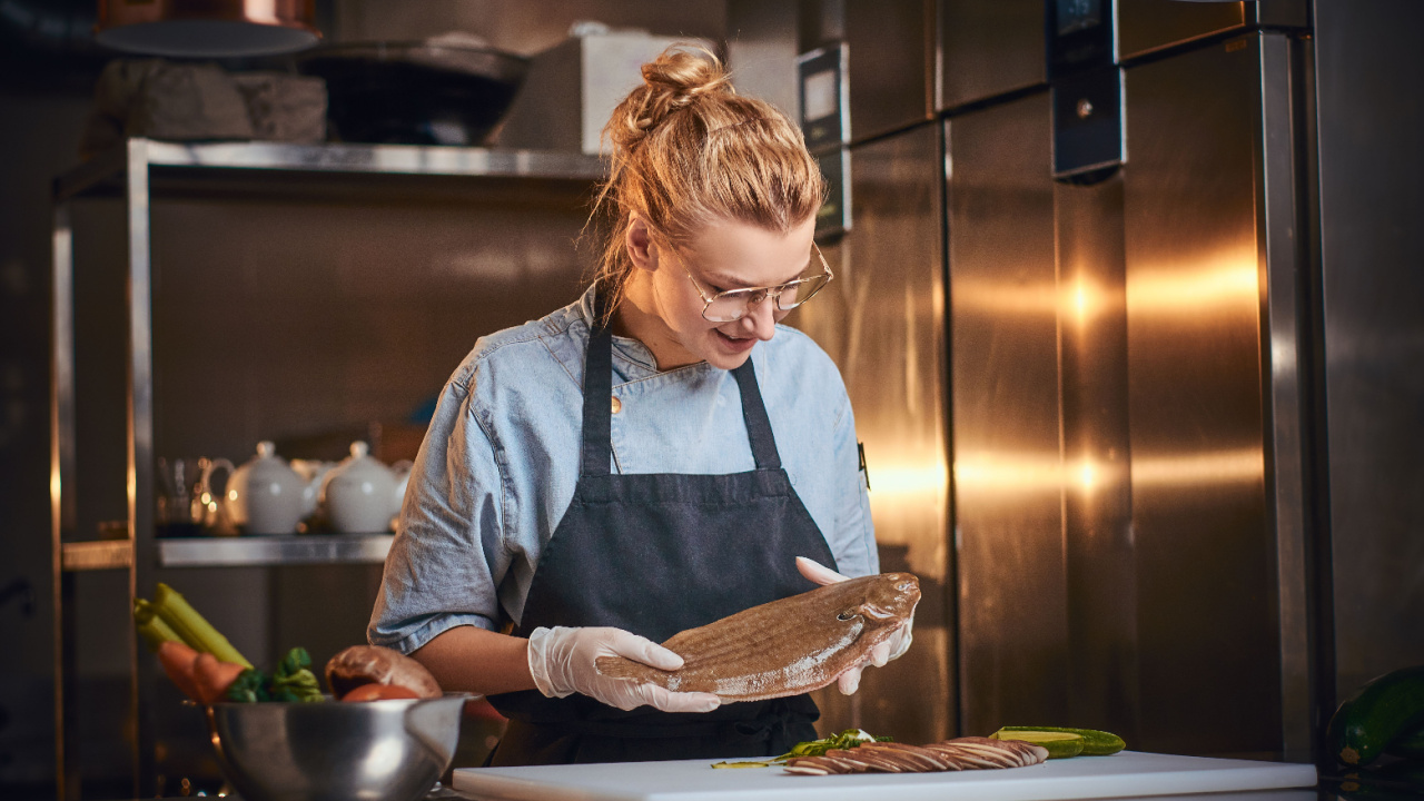 Female chef standing in a restaurant kitchen, cutting board with vegetables, holding a big fish, wearing apron and denim shirt