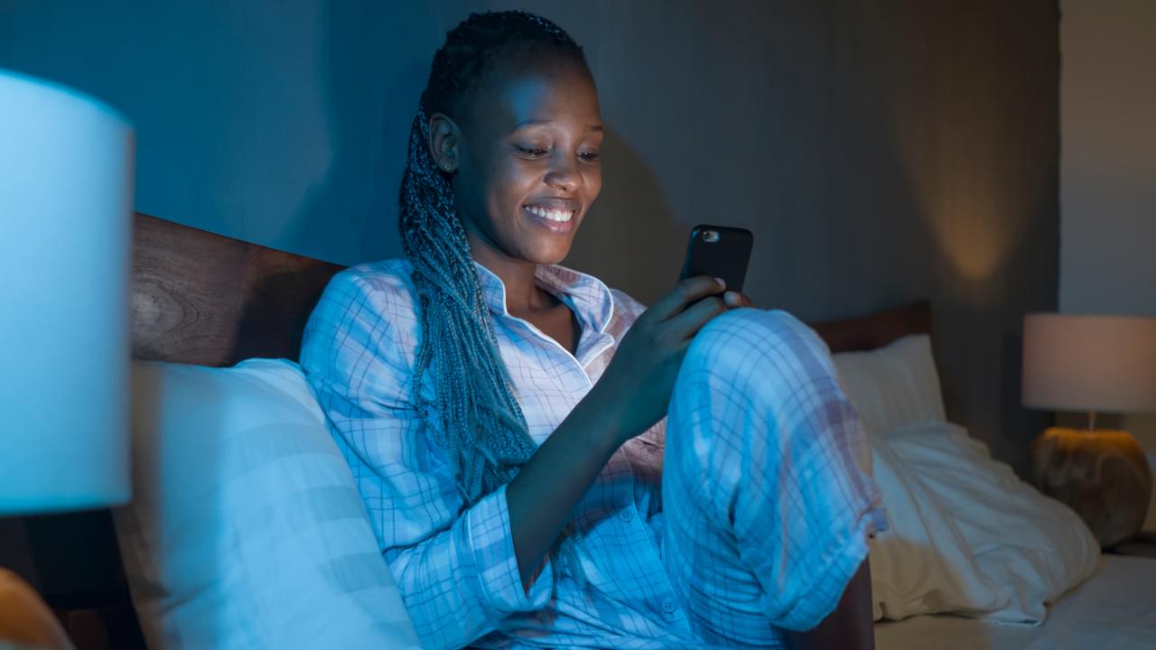 Happy African American millennial woman as social media addict - night lifestyle portrait of young beautiful and cool black girl texting or online dating on mobile phone in bed smiling cheerful