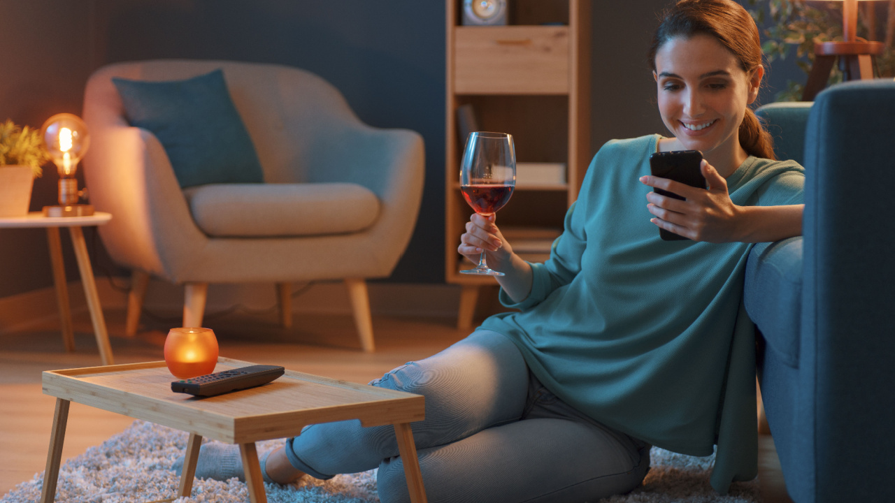 Happy woman relaxing at home couch, drinking a glass of wine, non alcohol and connecting with her smartphone