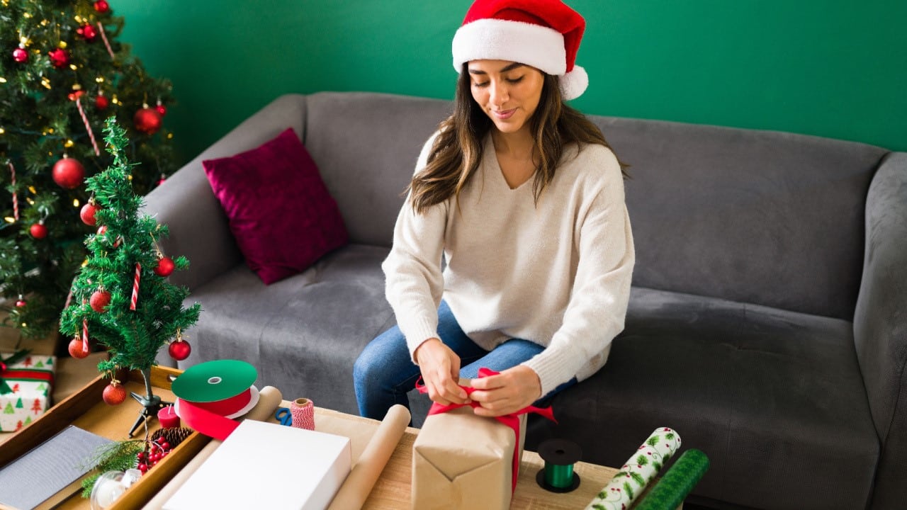 Happy young woman using wrapping paper and preparing her christmas gifts and envelopes before celebrating a party 