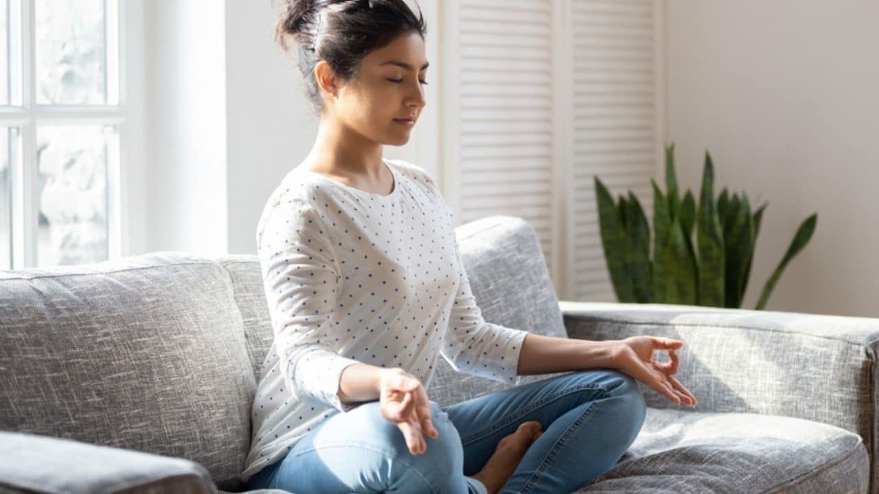 Indian female sitting on couch lotus pose put hands on lap folded fingers closed eyes enjoy meditation do yoga exercise at home. No stress, healthy habit, mindfulness lifestyle, anxiety relief concept