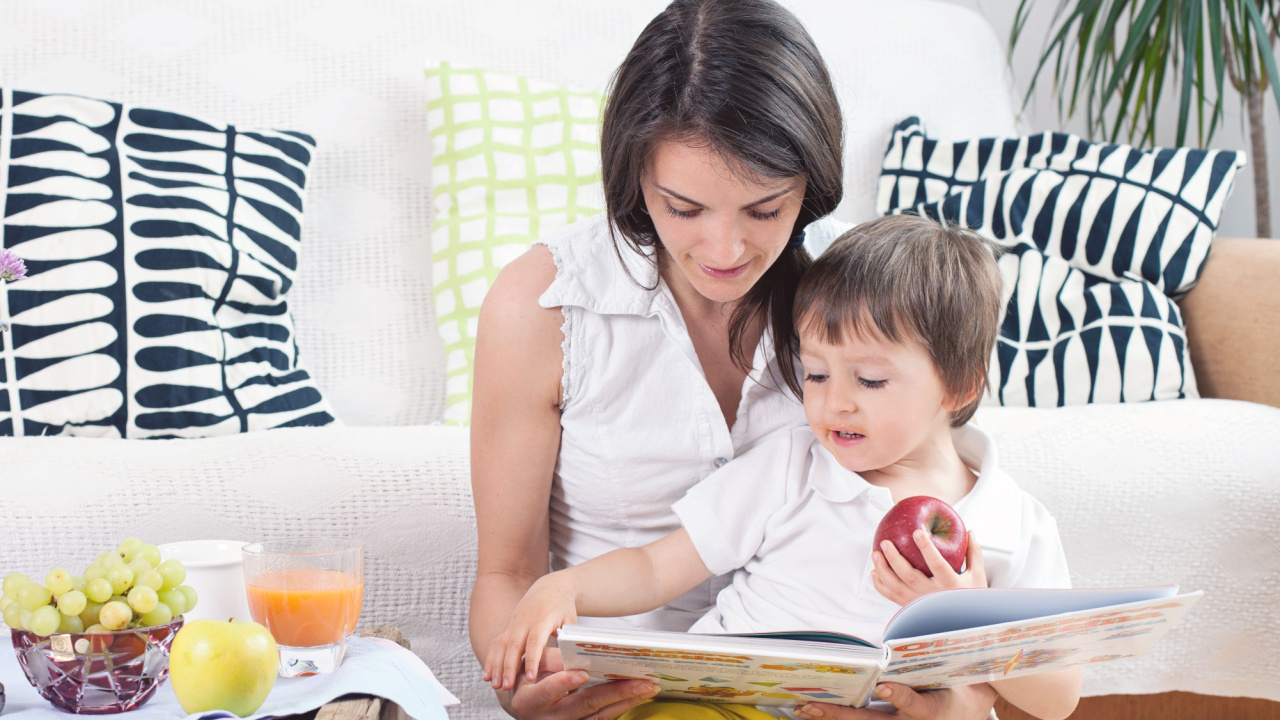 Mother and child, reading a book and eating fruits at home on a couch