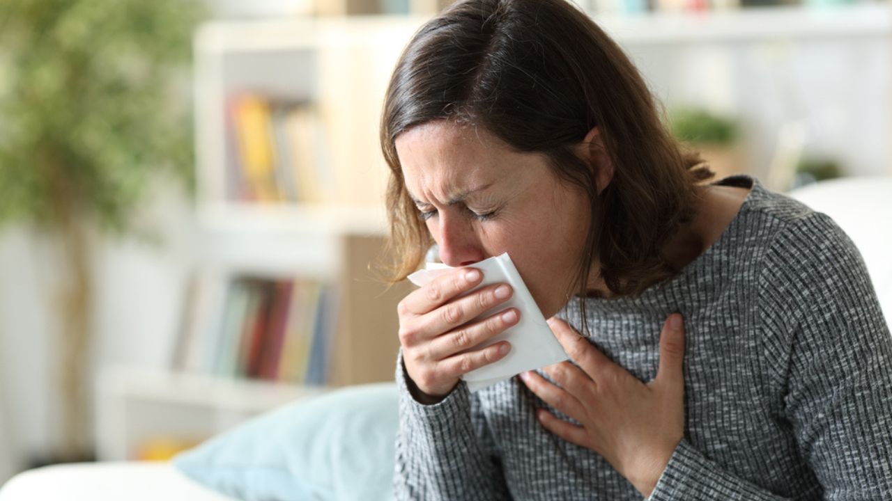 Sick adult woman coughing covering mouth with tissue sitting on a couch at home