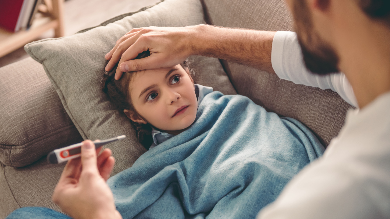 Sick little girl covered in blanket lying on couch, father taking temperature, flu