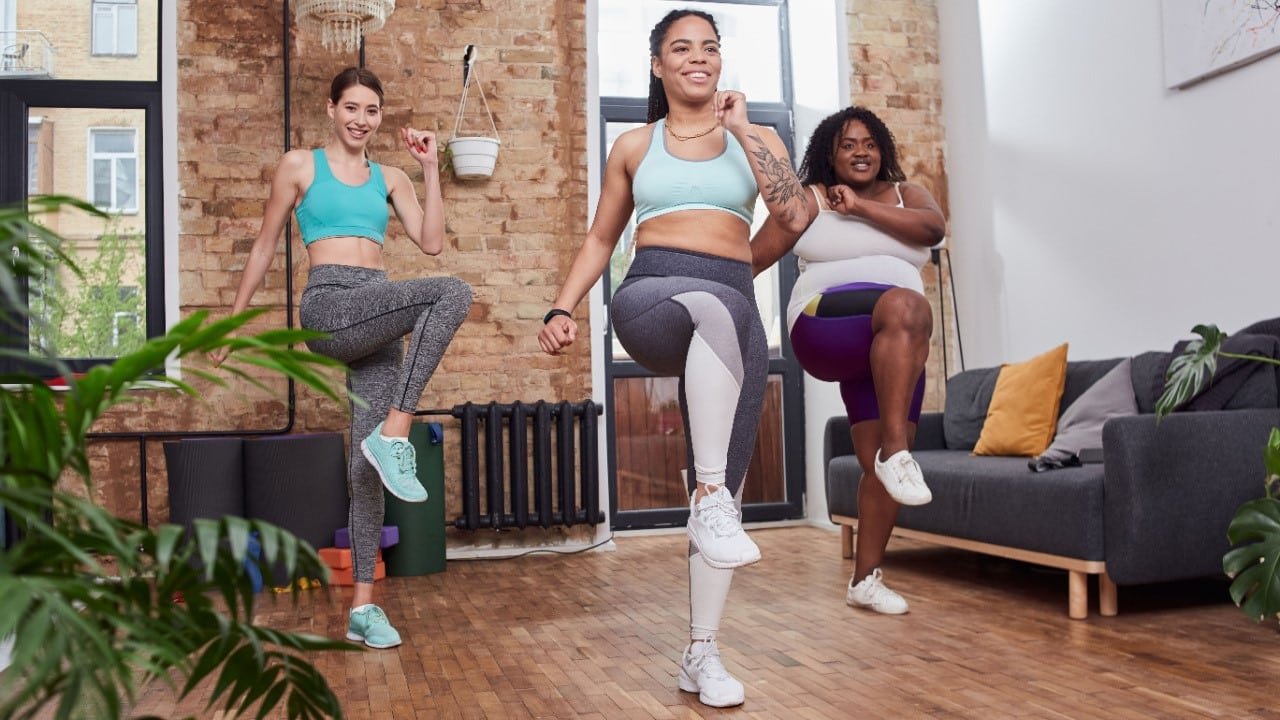 Smiling females doing high knees workout at home together making steps on place and rising their knees high