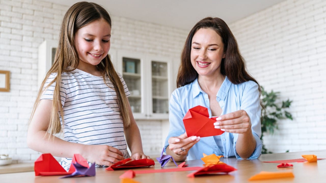 Smiling mother and daughter creating origami out of red paper in shape of heart, hobby activity