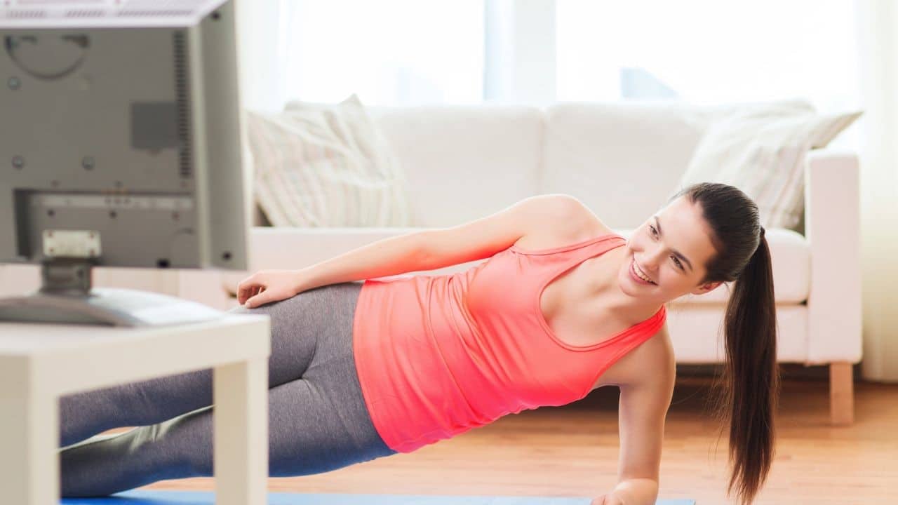 Woman in workout clothes doing a side plank while watching a workout video on TV, emphasizing an easy indoor exercise to boost step count.