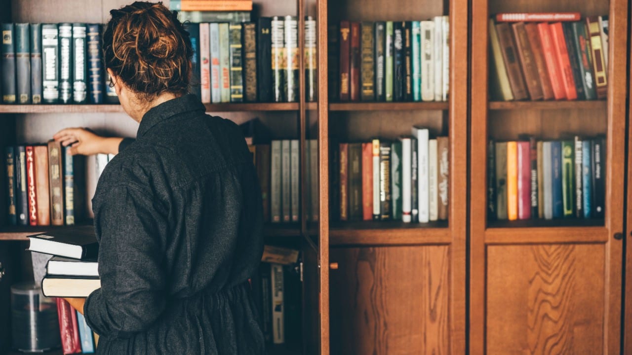 Young woman studying in the home library, picking books from the bookshelf