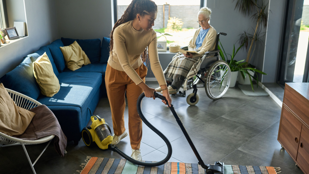 Youthful girl with vacuum cleaner helping her grandmother with disability